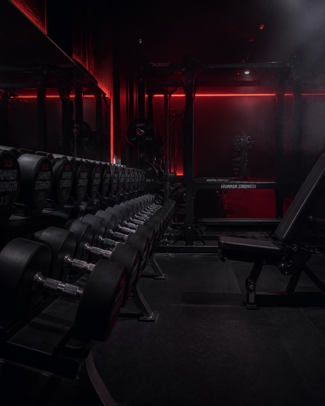Dark premium GYM BUDDY training floor with dumbbell racks under red accent lighting